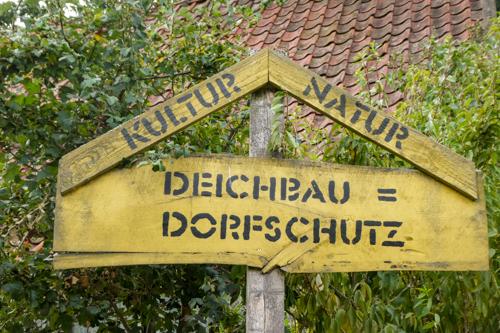 gelbes Holzschild mit Schriftzügen KULTUR NATUR und DEISCHBAU = DORTSCHUTZ